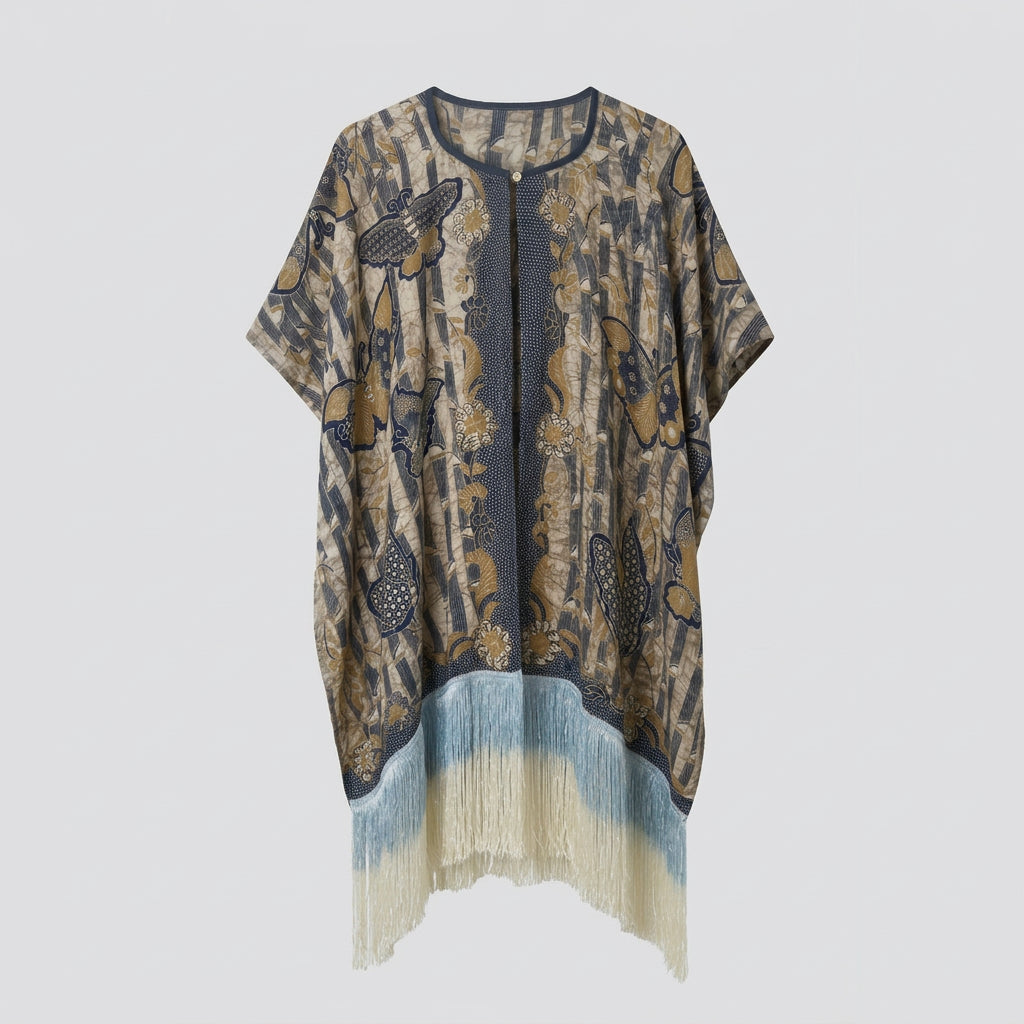 A loose, short-sleeve kaftan-style top hanging on a white hanger against a plain light wall. The garment has a Batik pattern butterfly and bamboo in beige, brown, black, and muted blue-gray, with a blue-gray gradient and fringe trim along the curved hem.