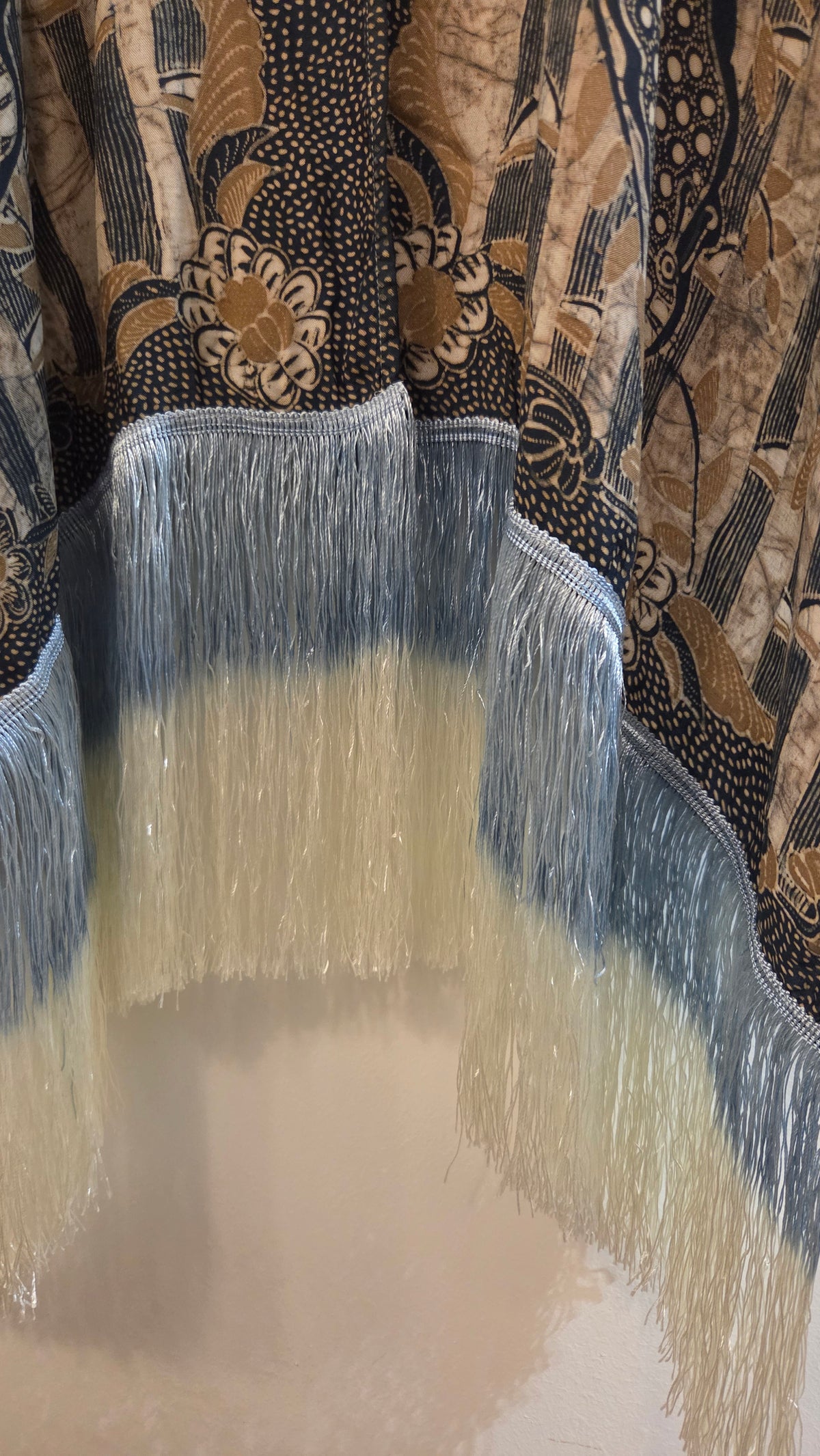 Close-up of a patterned cape with fringe, featuring a black, beige, and brown batik-style design with a blue-white fringe trim.