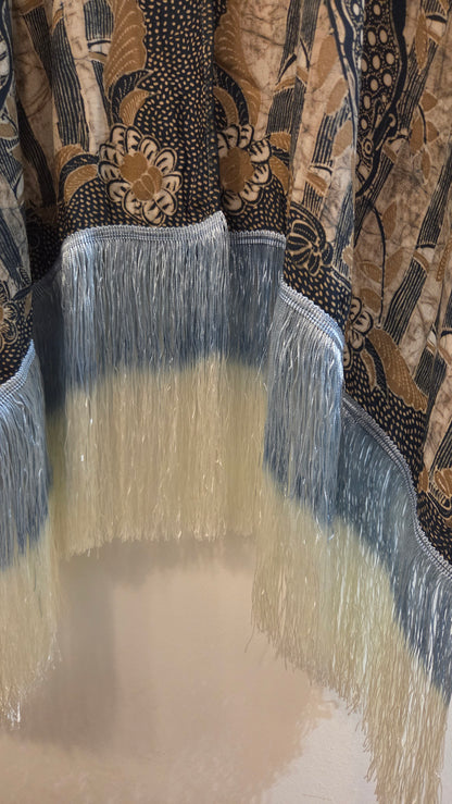 Close-up of a patterned cape with fringe, featuring a black, beige, and brown batik-style design with a blue-white fringe trim.
